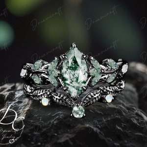 May include: A black ring set with a large, pear-shaped green gemstone surrounded by smaller green gemstones and opal accents. The ring features a delicate, leafy design.