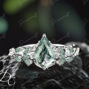 Puede incluir: Un anillo de plata con una piedra preciosa de ágata verde musgo en un engaste triangular. El anillo está adornado con piedras preciosas verdes más pequeñas y ramas de plata intrincadas.