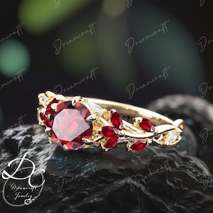 Könnte beinhalten: Ein goldener Ring mit einem großen roten Edelstein, der von kleineren roten Edelsteinen und weißen Akzenten umgeben ist. Der Ring hat ein zartes florales Design mit Blättern und Ranken.
