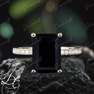 Könnte beinhalten: Ein schwarzer Smaragdschliff-Verlobungsring mit einer Pavé-Diamant-Bandfassung in 14 Karat Gold. Der Ring liegt auf einem dunkelgrauen Stein.