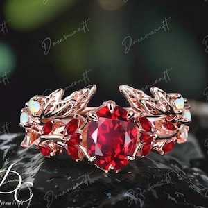 Peut inclure: Bague en or rose ornée d'une grosse pierre rouge ronde au centre, entourée de petites pierres rouges et de détails en forme de feuilles. La bague comporte également de petites pierres blanches. La bague est posée sur un fond sombre et texturé.