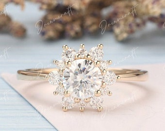 May include: A gold engagement ring with a round center stone surrounded by a halo of smaller stones. The ring is set on a white background.