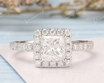 May include: A square-shaped diamond engagement ring with a halo setting. The ring is set in white gold and features a row of smaller diamonds around the band.