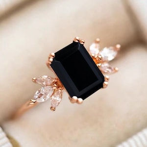 K&ouml;nnte beinhalten: Ein Ring aus Ros&eacute;gold mit einem gro&szlig;en, rechteckigen, schwarz gef&auml;rbten Stein im Smaragdschliff. Das Band ist auf beiden Seiten mit einer Gruppe von drei klaren, wei&szlig;en Steinen im Marquiseschliff verziert, die wie Bl&auml;tter angeordnet sind.