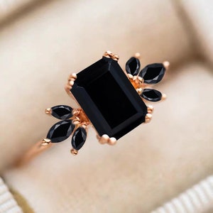 K&ouml;nnte beinhalten: Ein Ring aus Ros&eacute;gold mit einem gro&szlig;en, schwarz gef&auml;rbten Stein im Smaragdschliff, der auf jeder Seite von drei schwarzen Steinen im Marquiseschliff flankiert wird. Die Fassung besteht aus warmfarbigem Metall mit kleinen Krappen.