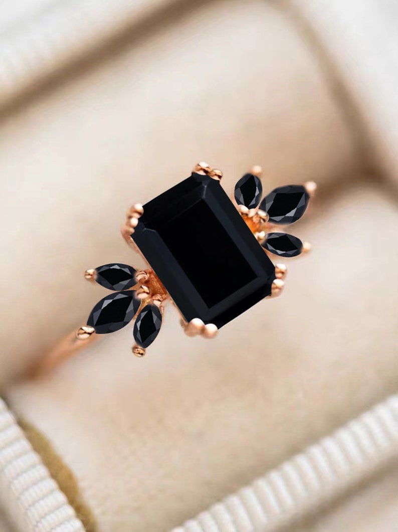 K&ouml;nnte beinhalten: Ein Ring aus Ros&eacute;gold mit einem gro&szlig;en, schwarz gef&auml;rbten Stein im Smaragdschliff, der auf jeder Seite von drei schwarzen Steinen im Marquiseschliff flankiert wird. Die Fassung besteht aus warmfarbigem Metall mit kleinen Krappen.