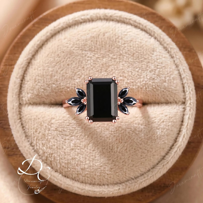 K&ouml;nnte beinhalten: Ein Ring aus Ros&eacute;gold mit einem gro&szlig;en, schwarz gef&auml;rbten Stein im Smaragdschliff, der auf jeder Seite von drei schwarzen Steinen im Marquiseschliff flankiert wird. Der Ring hat eine Krappenfassung, eine polierte Metallschiene und dunkle Edelsteine.