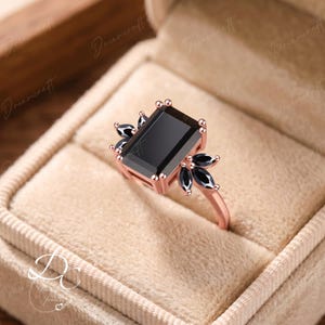 K&ouml;nnte beinhalten: Ein Ring aus Ros&eacute;gold mit einem gro&szlig;en, rechteckigen, schwarzfarbenen Mittelstein im Smaragdschliff, der an jeder Seite von drei kleineren, schwarzfarbenen Steinen im Marquiseschliff in einer Krappenfassung flankiert wird.