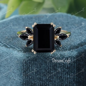 K&ouml;nnte beinhalten: Ein goldener Ring mit einem gro&szlig;en, smaragdgeschliffenen schwarzen Mittelstein, der auf jeder Seite von drei markisengeschliffenen schwarzen Steinen flankiert wird. Die Fassung hat kleine Krappen. Das Wort DreamCraft ist als Wasserzeichen sichtbar.