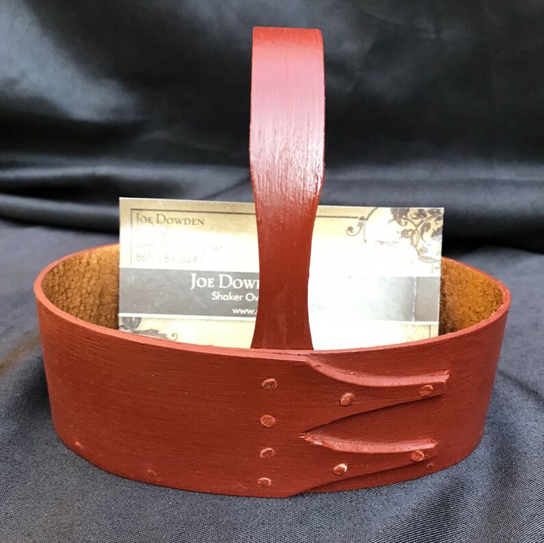 May include: A red wooden basket with a leather handle and a business card inside. The basket has a carved design and metal rivets. The business card reads "Joe Dowden, Shaker Oven, www.joedowden.com".