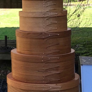 May include: A vertical stack of nine round wooden boxes of varying sizes, each featuring a light brown natural wood grain finish and a distinctive overlapping finger-jointed seam secured with small metal pins along the side of the container.
