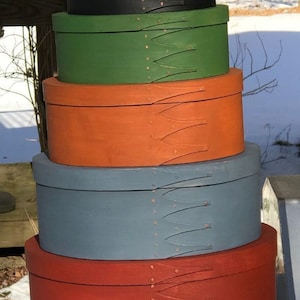 May include: A stack of eight oval wooden boxes, painted in various colors, including green, orange, blue, red, and black. The boxes are stacked in order of size, with the largest box at the bottom and the smallest box at the top. Each box has a decorative design of leather straps and copper rivets.