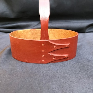 May include: A small, oval-shaped wooden basket with a red painted finish and a wooden handle. The basket has a natural wood interior.