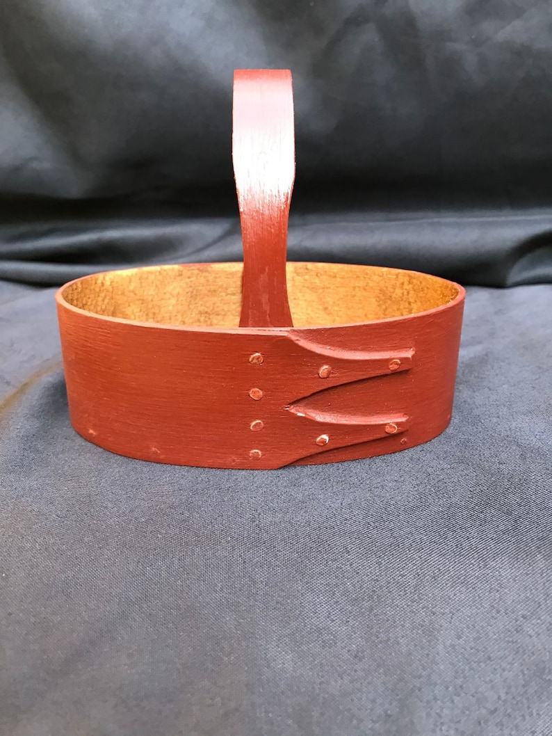 May include: A small, oval-shaped wooden basket with a red painted finish and a wooden handle. The basket has a natural wood interior.