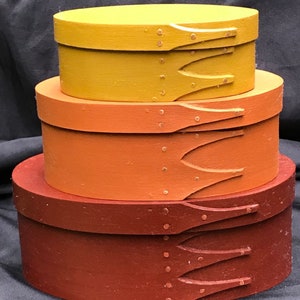 May include: Three nesting wooden boxes in shades of brown, orange, and yellow. The boxes are oval shaped and have a decorative carved design on the front.