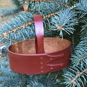May include: A small, oval-shaped, red wooden basket with a brown leather handle. The basket is hanging from a branch of a blue spruce tree.
