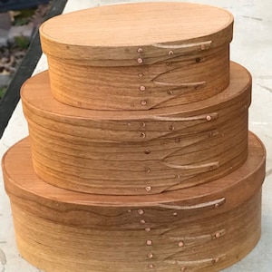 May include: Three wooden oval boxes with lids, nested together. The boxes are made of light brown wood with a natural grain pattern. The lids are secured with copper tacks.