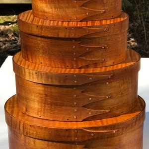 May include: Four wooden oval boxes stacked on top of each other. The boxes are made of light brown wood with a reddish-brown stain. The boxes are decorated with copper tacks and have a decorative design on the sides.
