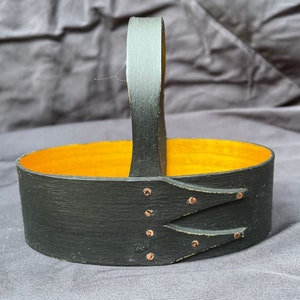 May include: A black oval wooden basket with a handle. The interior is painted a bright yellow-orange. The basket has decorative copper-colored rivets and a rustic, handcrafted appearance.