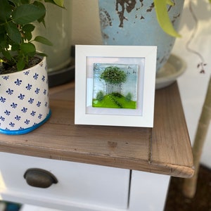 May include: A white framed glass art piece with a green landscape scene featuring a cluster of trees, a winding path, and a few birds in flight.
