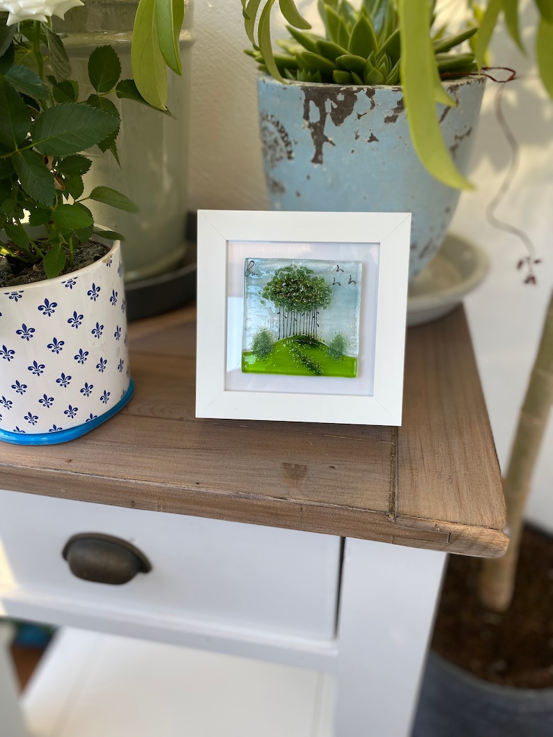 May include: A white framed glass art piece with a green landscape scene featuring a cluster of trees, a winding path, and a few birds in flight.