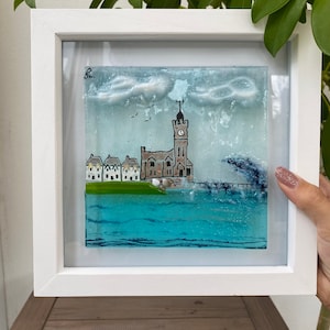 May include: A framed glass artwork depicting a seaside town with a clock tower, houses, and a pier. The sky is blue with white clouds and there are birds flying overhead. The water is blue and the waves are crashing against the shore.