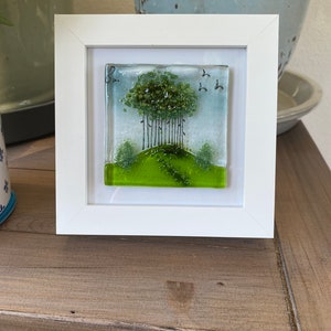 May include: A framed glass art piece depicting a green tree with a green grassy hill and a blue sky with birds flying above. The frame is white.