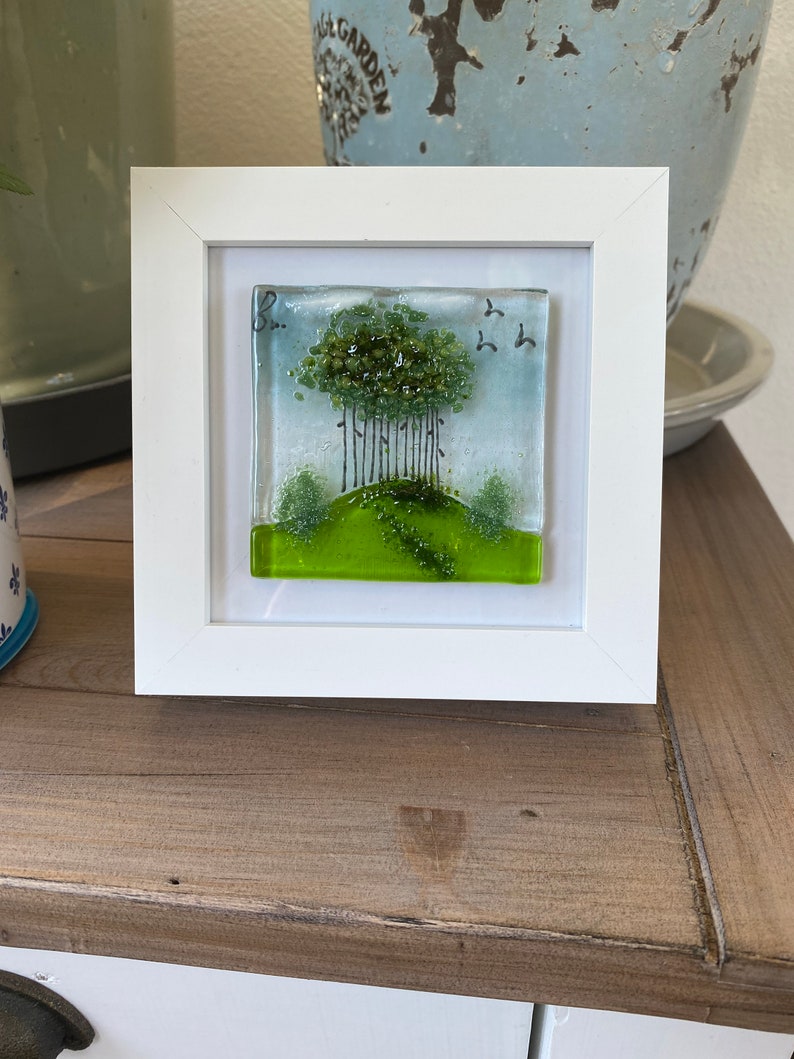 May include: A framed glass art piece depicting a green tree with a green grassy hill and a blue sky with birds flying above. The frame is white.