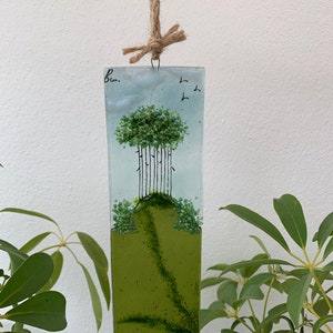 May include: A rectangular glass art piece depicting a green and blue landscape scene. The piece features a small hill with a line of trees on top, and a few birds flying in the sky. The piece is hanging from a piece of twine.