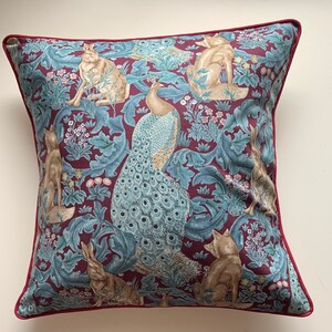 May include: Square decorative pillow with a burgundy background and a teal and beige pattern featuring foxes, rabbits, a peacock, and floral elements. The pillow has a burgundy trim.