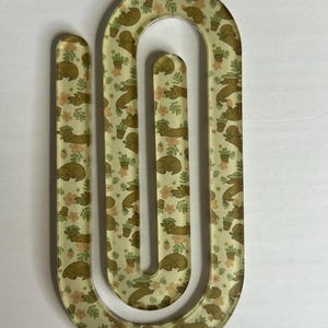 May include: A clear, U-shaped paperclip with a repeating pattern of sleeping dachshunds, potted plants, and small flowers in shades of brown, green, and peach. The paperclip is made of a transparent material.