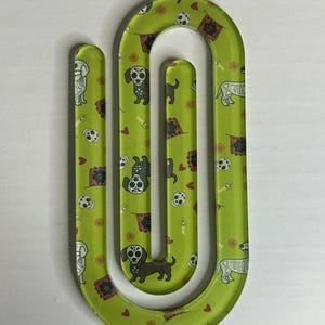 May include: A green, U-shaped paper clip with a repeating pattern of dachshunds, soccer balls, and decorative flags. The dachshunds are decorated with sugar skull designs. The paper clip is made of a clear, glossy material.