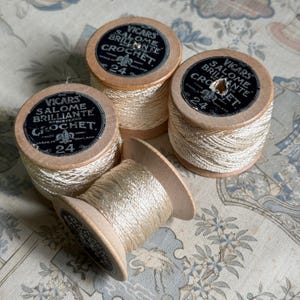 May include: Four wooden spools of cream-coloured crochet thread. Each spool features a black circular label with the text VICARS SALOME BRILLIANTE CROCHET, the number 24, and a fleur-de-lis emblem. The thread is wound tightly around the light wood.