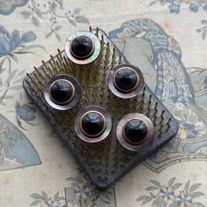 May include: Five black buttons with iridescent mother-of-pearl rims and gold-coloured metal accents, arranged on a rectangular floral pin frog. The pin frog is dark grey with numerous brass pins.