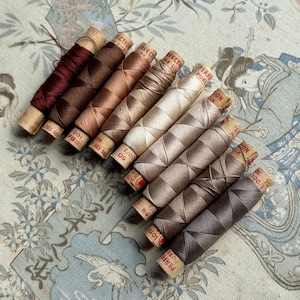 May include: A selection of silk thread spools in shades of brown, beige, and burgundy. The spools are arranged diagonally, displaying the different colours and textures of the thread. The spools are labelled with text and measurements.