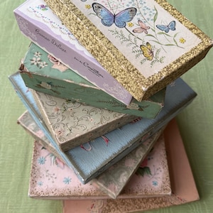 May include: A stack of rectangular boxes with decorative covers. The top box is white with a gold border, blue and yellow butterflies, and the text Cameo Deluxe from the Coronation Collection. Below are boxes in shades of green, blue, and pink with floral patterns.