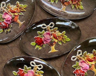 Könnte beinhalten: Mehrere ovale Glasdekorationen. Jedes Stück zeigt ein Blumenarrangement in einem Korb mit roten, rosa und gelben Blumen. Das Glas hat eine dunkle, reflektierende Oberfläche, und die floralen Details sind erhaben und metallisch.