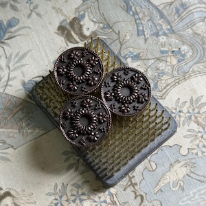 May include: Three circular dark brown buttons featuring a detailed embossed pattern with a central sunburst design surrounded by small leaf-like motifs. The buttons are arranged on a rectangular metal pin holder with numerous thin vertical spikes.