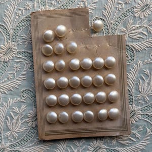 May include: A collection of round, off-white buttons with a smooth, pearlescent finish, arranged in rows on a rectangular tan cardboard display card. The card features thin, dark brown border lines and the text Made in Czechoslovakia at the bottom.