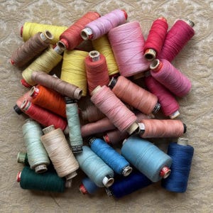 May include: A collection of assorted sewing thread spools in various sizes and colours, including shades of pink, red, orange, yellow, blue, green, and brown. The spools feature visible thread winding and cardboard cores with printed labels.