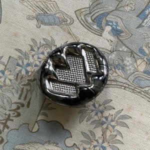 May include: A glossy, dark grey button with a textured, geometric pattern. The button is circular and has a reflective surface. The button is set against a floral patterned fabric.