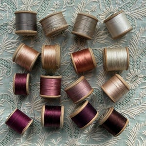 May include: A collection of fifteen wooden spools of thread in various shades of brown, beige, copper, and deep purple. The spools are arranged in a grid-like pattern, showcasing a gradient of neutral and warm metallic-toned sewing threads.