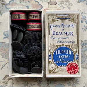 May include: A box of assorted black buttons with different patterns and textures. Beside it is a vintage box with French text, including "GRAND MAGASINS à RÉAUMUR Comptoir - Mercerie" and "FIL DE LIN EXTRA en Pelote".