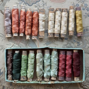May include: A collection of twenty-four vintage wooden spools of Lotus Button Twist thread in various colours, including shades of brown, green, red, yellow, and cream. Eleven spools are arranged in a light blue cardboard box, while thirteen are loose.