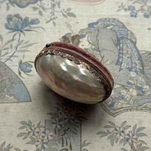 May include: An oval shell box with an iridescent surface, dark brown trim, and a purple velvet interior. The shell has a pearlescent sheen. The box is closed with a small, dark bead. The box is on a light blue and white floral patterned fabric.