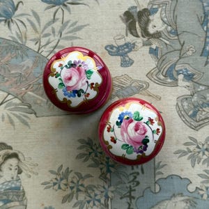 May include: Two round, deep red trinket boxes with white lids. Each lid features a hand-painted design of a central pink rose surrounded by small blue and purple flowers, accented with gold-coloured filigree detailing along the white edges.