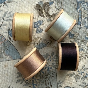 May include: Four thread spools in yellow, pale green, bronze, and dark brown. The thread is wound on wooden spools, set against a patterned fabric background. The spools are arranged in a circular pattern.
