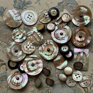 May include: A collection of assorted buttons in various sizes and shapes, primarily crafted from iridescent mother-of-pearl with a lustrous finish. The set includes round, flat, and rectangular pieces, some featuring metal centres or four-hole designs.