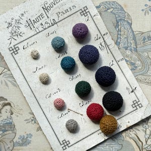May include: A selection of colourful, crocheted balls in various sizes and shades, including purple, blue, green, red, and gold, are arranged on a vintage paper. The paper, marked with "HAUTE NOUVELLE PARIS", has handwritten numbers and text.