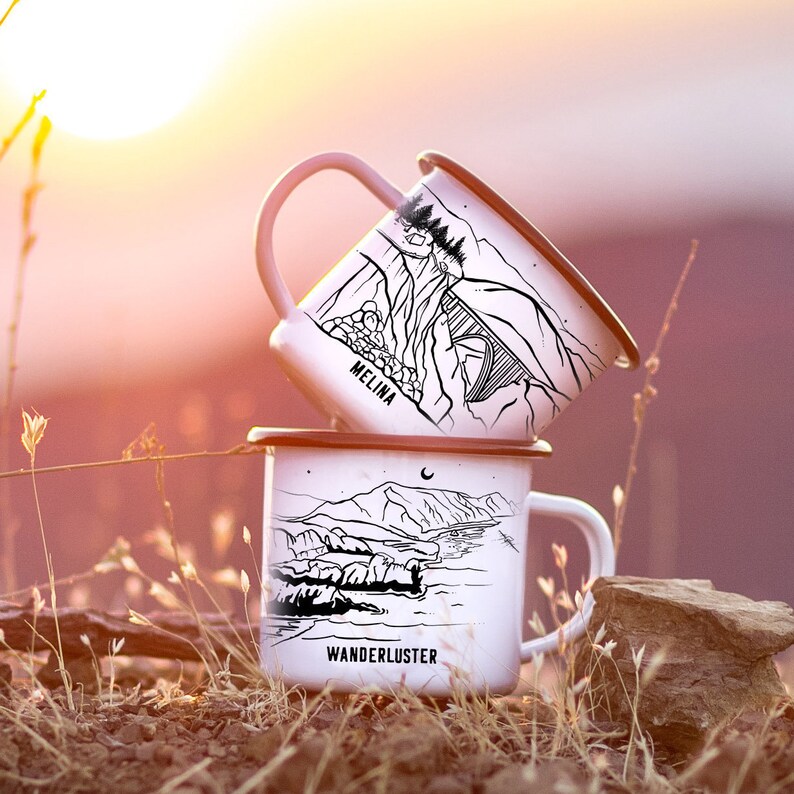 K&ouml;nnte beinhalten: Zwei wei&szlig;e Emailletassen mit schwarzen Illustrationen. Die obere Tasse zeigt eine Berglandschaft mit dem Wort "MELINA" und die untere Tasse zeigt eine Strandlandschaft mit dem Wort "WANDERLUSTER".
